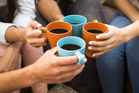 Four Individuals sitting in a circle hold coffee mugs together as they make a toast.