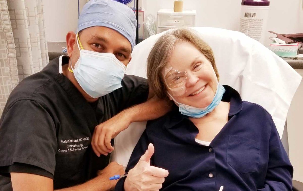 eye surgeon in scrub cap and mask posing post-surgery next to female patient who has a thumbs up