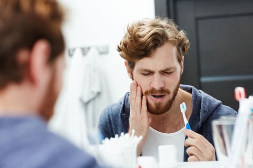 Man holding jaw from tooth sensitivity looking in a bathroom mirror