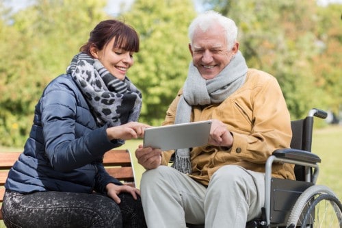 Young woman helping elderly man in wheelchair outdoors with tablet