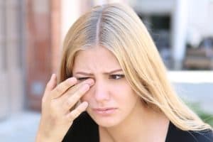 Blonde woman rubs her eye while outside due to dry eye symptoms.