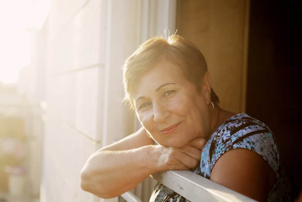 Senior woman smiles in the sunshine while looking outside of her window